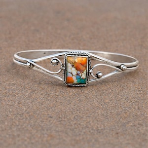 May include: A silver-toned bracelet with a rectangular, multi-colored stone at the center. The stone features shades of orange, white, and turquoise. The bracelet has a delicate, open design with decorative swirls and small ball accents.
