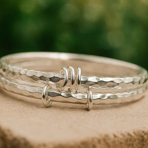 Könnte beinhalten: Drei silberne Armreifen mit gehämmerten Strukturen, verbunden durch kleine, kreisförmige silberne Akzente. Der Schmuck wird auf einer hellbraunen Oberfläche vor einem verschwommenen grünen Hintergrund präsentiert.