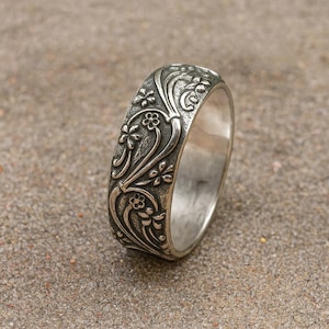 Puede incluir: Un anillo de plata con un patrón floral grabado. El anillo presenta un diseño repetido de flores y vides alrededor de la banda. El anillo se muestra sobre una superficie neutra.
