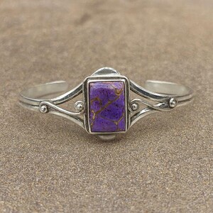 May include: Silver cuff bracelet with a rectangular, purple stone with gold veining. The bracelet has an open design with decorative swirls and small silver beads. A piece of jewellery.