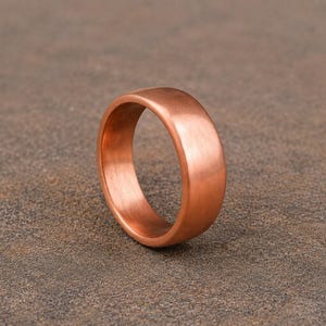 Puede incluir: Un anillo de cobre macizo con un acabado liso y pulido y un perfil redondeado. La banda presenta un tono marrón rojizo metálico cálido con sutiles variaciones de tono en toda su superficie, mostrando un diseño circular simple.