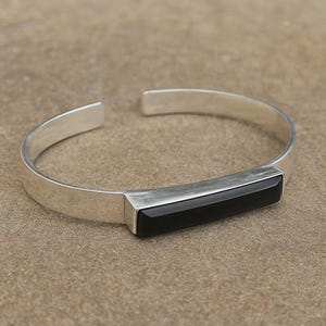 Op de afbeelding: Een zilveren manchetarmband met een rechthoekige zwarte onyxsteen in het midden. De armband heeft een eenvoudig, modern ontwerp met een gepolijste afwerking. Het open ontwerp maakt het gemakkelijk om te dragen.