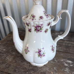 May include: White porcelain teapot with a lid, decorated with purple floral patterns and gold trim. The teapot has a curved spout and a handle. The lid has a finial. The teapot is sitting on a wooden surface.
