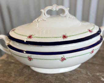May include: A white porcelain soup tureen with a lid, featuring a navy blue band and delicate floral and green vine detailing. The tureen has two handles and a decorative handle on the lid. A classic piece for serving soups and stews.