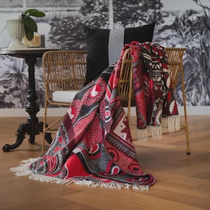 Könnte beinhalten: Ein gewebter Rattanstuhl mit einer rot, schwarz und weiß gemusterten Decke. Die Decke hat einen Fransenbesatz. Ein schwarzes Kissen mit grauem Streifen befindet sich hinter der Decke. Links steht ein kleiner schwarzer Tisch.