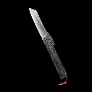 May include: A handcrafted folding knife with a textured, dark metal handle and a silver blade. The blade has a unique, slightly curved shape. A small leather cord is attached to the base of the handle. The knife is set against a black background.
