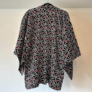 以下が含まれることがあります： 白線とピンクのドットの繰り返し模様の黒い着物。 黒いハンガーに白い壁を背景に吊るされています。 袖は広く、生地は軽量に見えます。