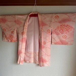 以下が含まれることがあります： 白と濃いピンクの花柄があしらわれた、絞り風のテクスチャーを持つ淡いピンク色の羽織。幅の広い袖とまっすぐな前開きが特徴で、中央には白い裏地が見えています。ハンガーに掛けられた状態の衣類です。