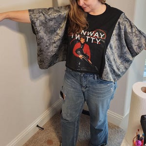 May include: Black crop top with a graphic of a man in a red jacket holding a guitar, with the text "Runway Kitty". The sleeves are made of gray paisley fabric. The model is wearing blue jeans.