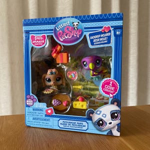 May include: A Littlest Pet Shop toy set in a blue box. The set includes two animal figures, accessories, and a backdrop. The box features a panda illustration and text in English and Spanish.