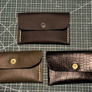 Peut inclure: Trois porte-cartes en cuir noir, vert olive et marron foncé. Chacun a une fermeture à rabat avec un bouton-pression ou un bouton et des coutures blanches. Les porte-cartes sont rectangulaires et semblent faits à la main.