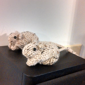 May include: Two knitted grey mice figurines on a black surface. The mice are sitting side by side, with the one on the right slightly larger than the one on the left.