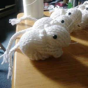 May include: A group of white crocheted mice with black eyes. The mice are arranged in a line, with their tails trailing behind them.