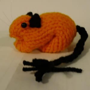 May include: A hand-knitted orange and black plush toy shaped like a guinea pig. The toy has a long, black tail and black eyes.