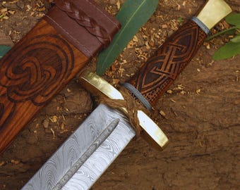 May include: A decorative sword with a patterned blade and a wooden handle featuring intricate carvings. The handle has brass accents and is wrapped with twine. A wooden sheath with a leather top is also visible.
