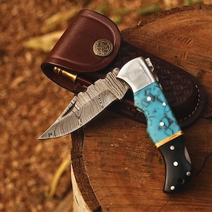 May include: A folding pocket knife with a Damascus steel blade, a turquoise and black handle with silver pins, and a brown leather sheath featuring a circular brass-colored button closure with a star emblem embossed on the front surface.