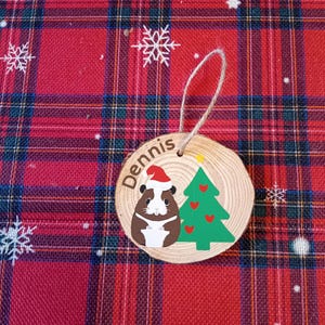 May include: A round wooden Christmas bauble featuring a brown and white guinea pig wearing a red Santa hat and a green Christmas tree with red hearts. The bauble is personalised with the name "Dennis".