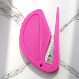K&ouml;nnte beinhalten: Ein leuchtend pinkfarbener Brief&ouml;ffner mit integrierter Klinge. Der &Ouml;ffner hat einen gebogenen Griff und einen spitzen Klingenschutz. Die Klinge ist silberfarben und hat zwei kleine L&ouml;cher. Das Design ist modern und elegant.