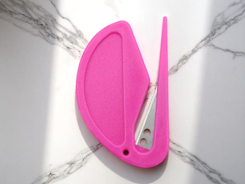 K&ouml;nnte beinhalten: Ein leuchtend pinkfarbener Brief&ouml;ffner mit integrierter Klinge. Der &Ouml;ffner hat einen gebogenen Griff und einen spitzen Klingenschutz. Die Klinge ist silberfarben und hat zwei kleine L&ouml;cher. Das Design ist modern und elegant.