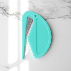 K&ouml;nnte beinhalten: Ein t&uuml;rkisfarbener Brief&ouml;ffner mit einer silbernen Klinge. Der &Ouml;ffner hat einen gebogenen Griff und ein abgerundetes Ende mit einem kleinen Loch in der N&auml;he der Oberseite. Das Design ist modern und minimalistisch.