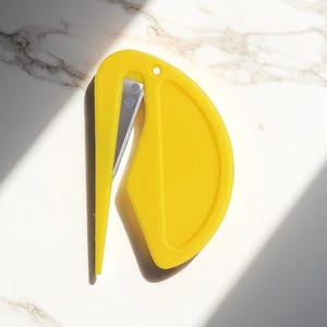 K&ouml;nnte beinhalten: Ein leuchtend gelber Brief&ouml;ffner mit geschwungenem Design und silberner Klinge. Der &Ouml;ffner hat oben ein kleines Loch und liegt vor einem wei&szlig;en Marmorhintergrund mit Schatten.