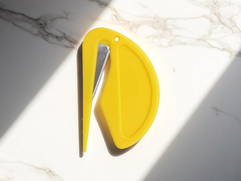 K&ouml;nnte beinhalten: Ein leuchtend gelber Brief&ouml;ffner mit geschwungenem Design und silberner Klinge. Der &Ouml;ffner hat oben ein kleines Loch und liegt vor einem wei&szlig;en Marmorhintergrund mit Schatten.