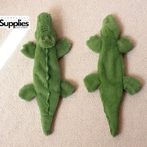 Könnte beinhalten: Zwei grüne Plüsch-Alligator-Hundespielzeuge. Die Spielzeuge liegen auf einer beigefarbenen Oberfläche. Die Alligatorspielzeuge haben ein strukturiertes, weiches Aussehen mit Kopf, vier Gliedmaßen und einem Schwanz. Die Spielzeuge sind für Haustiere konzipiert.