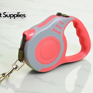 Op de afbeelding: Een intrekbare hondenriem in koraal en grijs. De riemhandgreep is koraal, met een grijze behuizing en een koraalrood midden. De riem heeft een verchroomde sluiting en een zwarte band. De tekst "PAWSOMESTUFF Pet Supplies" staat linksboven.