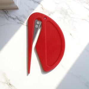 K&ouml;nnte beinhalten: Ein roter Brief&ouml;ffner mit gebogenem Griff und scharfer, silberner Klinge. Der &Ouml;ffner hat an einem Ende eine abgerundete, schildartige Form. Das Design ist schlicht und modern, geeignet zum &Ouml;ffnen von Umschl&auml;gen und Paketen.
