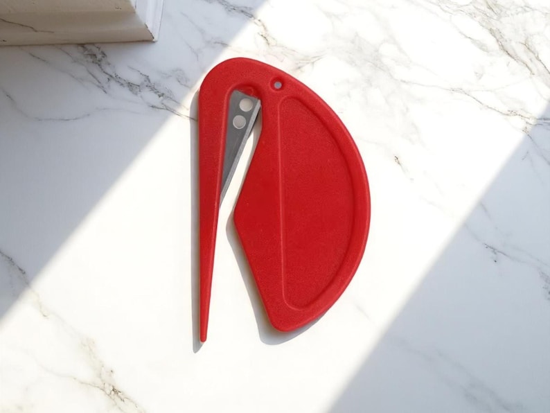 K&ouml;nnte beinhalten: Ein roter Brief&ouml;ffner mit gebogenem Griff und scharfer, silberner Klinge. Der &Ouml;ffner hat an einem Ende eine abgerundete, schildartige Form. Das Design ist schlicht und modern, geeignet zum &Ouml;ffnen von Umschl&auml;gen und Paketen.
