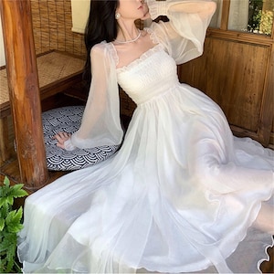 Puede incluir: Un vestido largo blanco con escote cuadrado y mangas largas transparentes. El corpiño está fruncido y la falda llega hasta el suelo. El vestido está hecho de una tela ligera y semitransparente, creando un aspecto etéreo. Adecuado para ocasiones especiales.