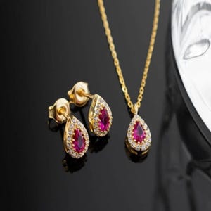 Op de afbeelding: Een goudkleurige sieradenset met een hangerketting en bijpassende oorstekers. Elk stuk heeft een druppelvormige, robijnrode middensteen omgeven door kleine, heldere accentstenen. De set wordt gepresenteerd op een zwart oppervlak.