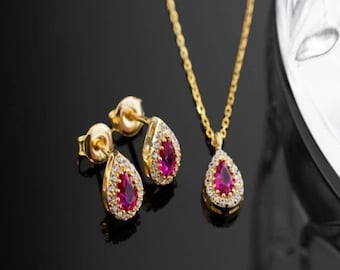 May include: A gold-toned jewelry set featuring a pendant necklace and matching stud earrings. Each piece has a teardrop-shaped, ruby-colored center stone surrounded by small, clear accent stones. The set is displayed on a black surface.