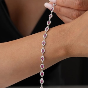 May include: A silver bracelet with a repeating pattern of teardrop-shaped, crimson gemstones, each encircled by smaller, clear stones. The bracelet is displayed against a dark backdrop.