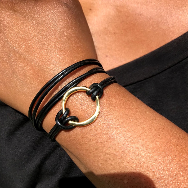 May include: A black leather bracelet with a gold circle charm. The bracelet is wrapped around a wrist.