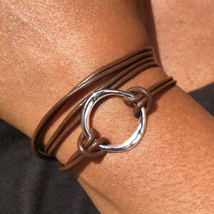 May include: A brown leather wrap bracelet with a silver circle charm. The bracelet is wrapped around a person's wrist.