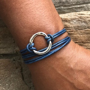 May include: A blue leather wrap bracelet with a silver circle charm. The bracelet is made of multiple strands of leather and has a simple, elegant design.