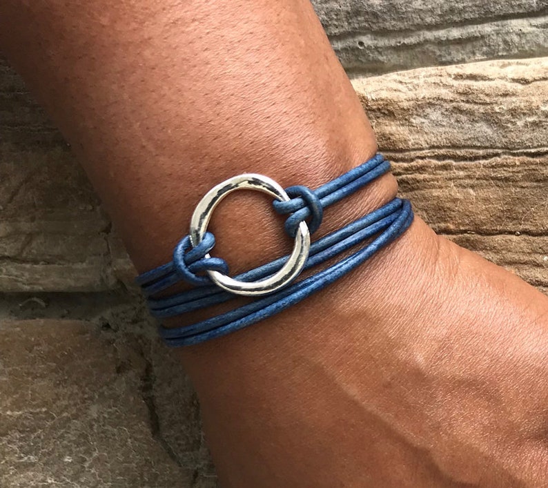 May include: A blue leather wrap bracelet with a silver circle charm. The bracelet is made of multiple strands of leather and has a simple, elegant design.