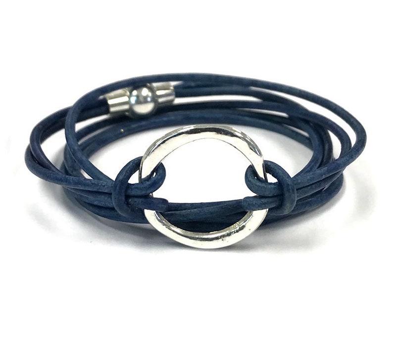 May include: A navy blue leather wrap bracelet with a silver circle charm. The bracelet is made of multiple strands of leather and has a magnetic clasp.