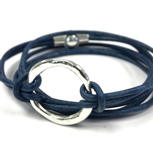 May include: A blue leather wrap bracelet with a silver circle charm. The bracelet is made of multiple strands of leather that are wrapped around the wrist and secured with a magnetic clasp.