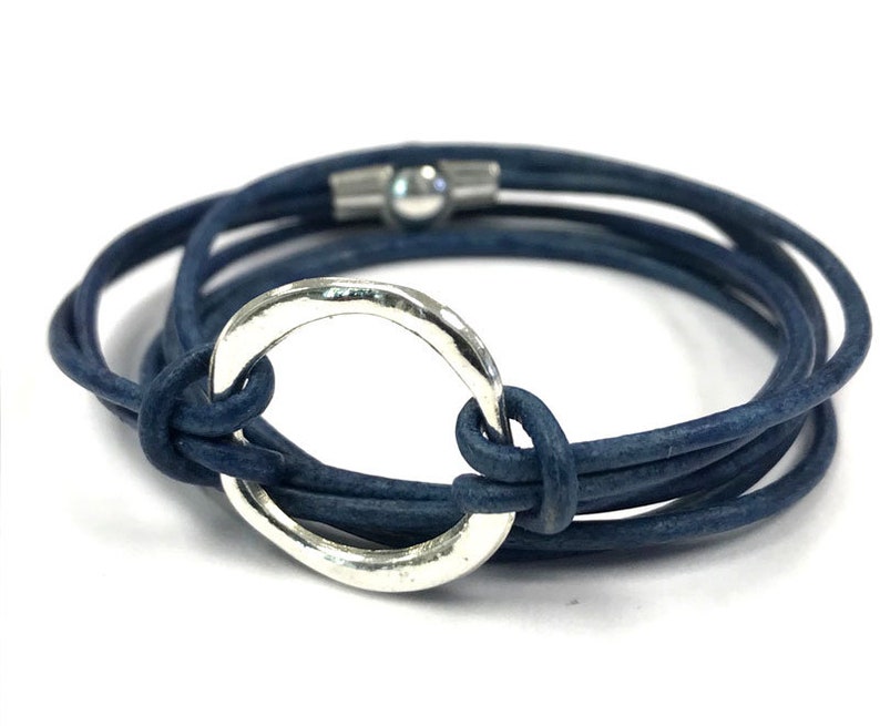 May include: A blue leather wrap bracelet with a silver circle charm. The bracelet is made of multiple strands of leather that are wrapped around the wrist and secured with a magnetic clasp.