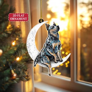 May include: A 2D flat ornament featuring a blue heeler dog sitting on a white crescent moon. The dog has a detailed painted appearance with shades of blue, black, and brown. The ornament hangs from a gold string and has the text "2D FLAT ORNAMENT".