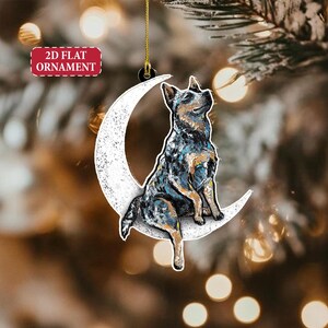 May include: A 2D flat ornament featuring a dog sitting on a crescent moon. The dog has a blue, black, and tan coat. The moon is white with a textured appearance. The ornament has a gold hanging string and the text "2D FLAT ORNAMENT".