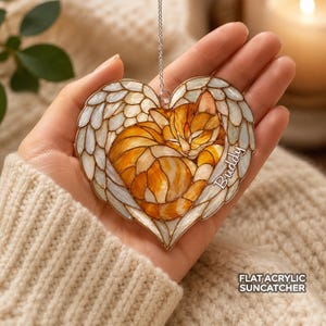 May include: A heart-shaped flat acrylic suncatcher featuring a stained glass style illustration of an orange tabby cat curled up inside white angel wings. The name Buddy is printed in white script on the right side of the heart ornament.