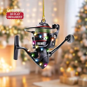 May include: A black and silver fishing reel ornament with green fishing line. The ornament has the word "NAME" on it and is decorated with colorful lights. The text "Custom text 2D FLAT ORNAMENT" is in the upper left corner.