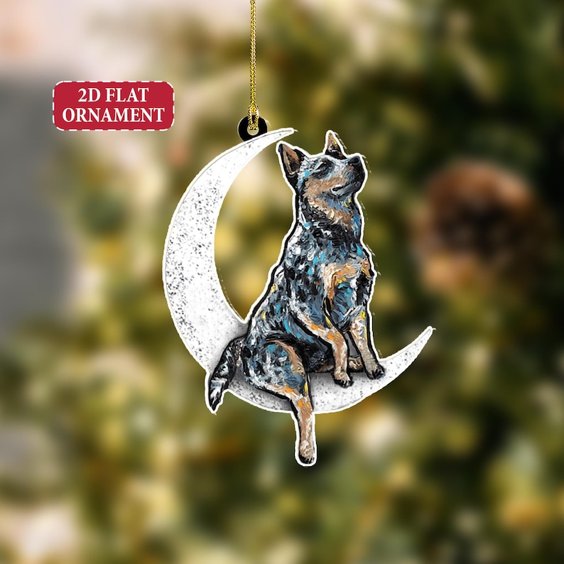 May include: A 2D flat ornament featuring a blue heeler dog sitting on a white crescent moon. The dog is painted with blue, black, and tan colors. The ornament has a gold string for hanging and a red label that says "2D FLAT ORNAMENT".