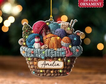 May include: A 2D flat ornament shaped like a basket filled with yarn balls, knitting needles, and a snowman. The basket is brown with a knitted border and the name "Amelia" on a label. The ornament has a gold string for hanging and the text "Custom text 2D FLAT ORNAMENT".