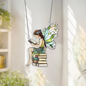 May include: A stained glass fairy reading a book, perched atop a stack of books. The fairy has brown hair, a green and yellow dress, and large, colorful wings. The piece hangs from a silver chain.
