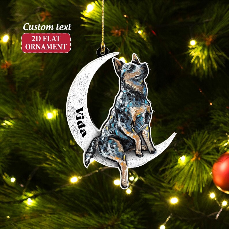 May include: A 2D flat ornament featuring a dog sitting on a crescent moon. The dog has blue, black, and brown markings. The moon is white with the name "Vida" printed on it. The ornament has the text "Custom text" and "2D FLAT ORNAMENT".