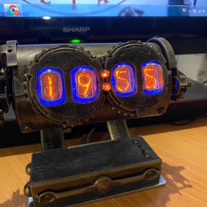 Pode incluir: Um relógio de tubos nixie com quatro visores numéricos laranja brilhantes, alojados numa caixa metálica escura e desgastada com molduras circulares dentadas. O dispositivo inclui duas pequenas luzes indicadoras âmbar e uma base retangular.